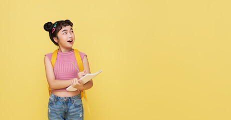 Young Asian kid girl thinking and writing something on notebook isolated on yellow background.