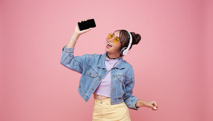 Happy beautiful Asian teen woman in yellow glasses listening music in headphones and dance on pink...