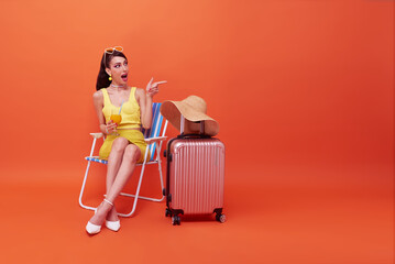 Beautiful woman relax sitting on a beach chair with suitcase pointing her finger in studio summer...