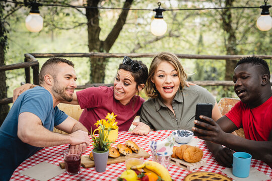 happy friends having breakfast in the backyard, young influencer using smartphone to create content on social media with photos of food, multiracial couples having fun at picnic outside