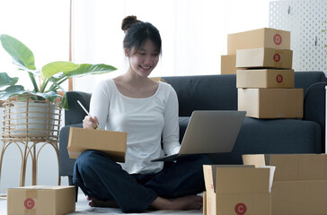 Fototapeta premium Smiling young Asian business owner woman prepare parcel box and standing check online orders of product for deliver to customer on laptop computer. Shopping Online concept.