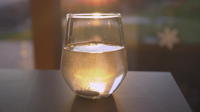 SLOW MOTION, CLOSE UP: Effervescent tablet dissolving in sunlit glass of water. Sparkling beverage for regaining health backlit with beautiful golden light. Seasonal home treatment for immunity boost.