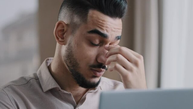 Exhausted young millennial businessman overworked male worker employee massaging dry irritable eyes feeling eyestrain after using computer tired hispanic man guy have pain headache bad vision problem