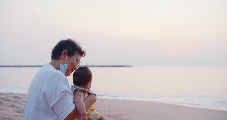 happiness Asian family grandmother is keeping holding cute adorable newborn baby infant girl on her arms relaxing at seaside tropical beach sunrise morning during holiday vacation in Thailand	