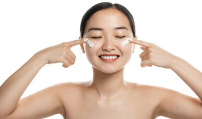 Woman applying skincare product on her face. Asian woman putting cosmetic cream in her face and smiling against white background