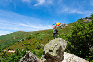 A woman with a backpack, perched on a rock on top of a mountain, arms raised holding a rainbow flag waving in the wind.