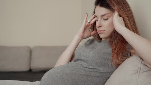 Young pregnant woman in gray clothes sitting on the couch, feeling unwell and having a headache.