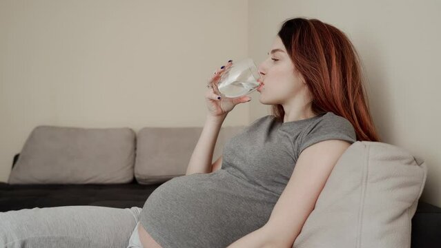 Young pregnant woman in gray clothes drinking water and taking pills.