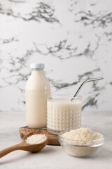 A bottle and a glass of rice milk with a bowl of rice and a wooden spoon on a wooden board on a light background