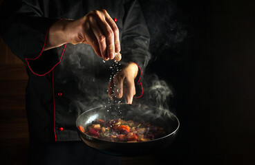 Professional chef adds salt to a steaming hot pan. Grande cuisine idea for a hotel with advertising space. - obrazy, fototapety, plakaty