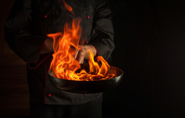 The chef cooking food in pan with fire flame on black background. Restaurant and hotel service...