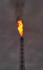 Vertical shot of a flaming gas torch in an oil refinery against a gloomy sky. Burning refinery torch contaminating the environment with smoke and causing pollution and climate change.