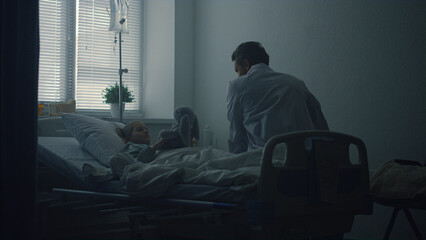 Doctor visiting little patient in clinic ward. Sick girl hug plush toy in bed.