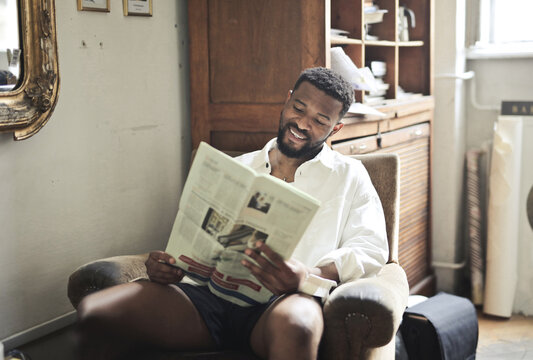 portrait of young man smiling while reading a newspaper sitting in an armchair
