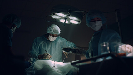Doctors team performing surgical operation in dark hospital operating room. - obrazy, fototapety, plakaty