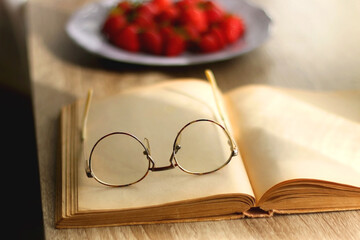 Lilac plate full of fresh strawberries, open book and reading glasses on wooden table. Selective focus.