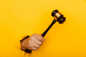 Male hand holding a wooden gavel through torn yellow background. Law and auction aconcept - obrazy, fototapety, plakaty