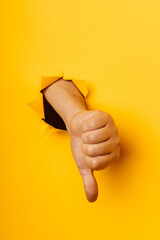 Hand showing a thumb down through ripped hole in yellow paper background. Concept of dislike and disapproval gesture. Vertical image