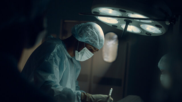 Focused Surgeon Performing Operation In Dark Sterile Hospital Operating Room.