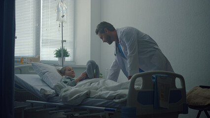 Friendly doctor talking little patient in ward. Modern operating room interior.