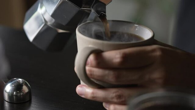 Barista pouring coffee from moka pot coffee maker to a coffee cup. Hand holding Italian classic moka pot pouring coffee.