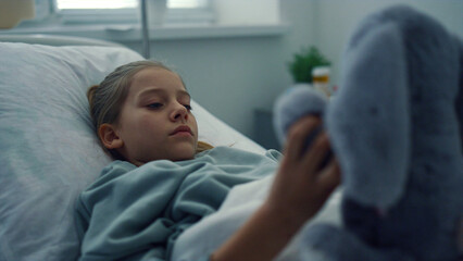 Upset girl lying hospital bed portrait. Sad kid playing with plush toy alone.