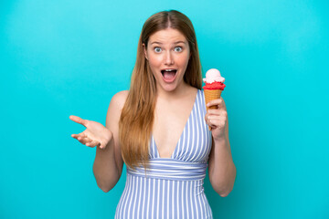 Young caucasian woman in swimsuit eating ice cream isolated on blue background with shocked facial expression