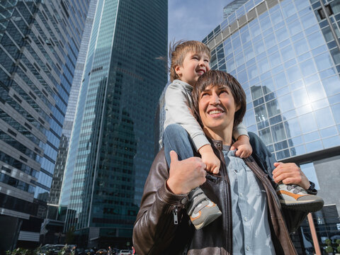 Little boy sits on father's shoulders among skyscrapers. Dad and son looks on glass walls of buildings. Future and modern technologies, life balance and family life in well keeps districts.