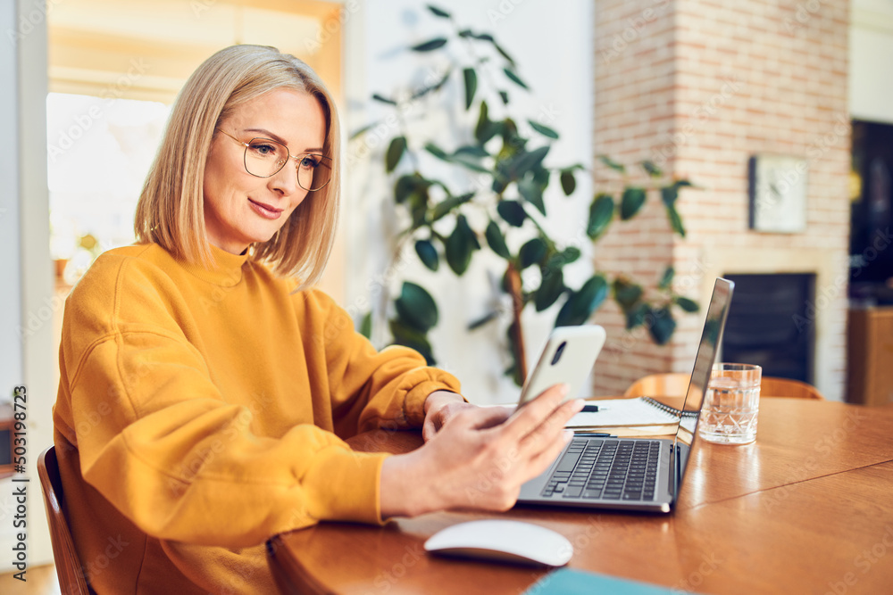 Wall mural mature woman with glasses looking on smartphone sitting at dinning table while working from home