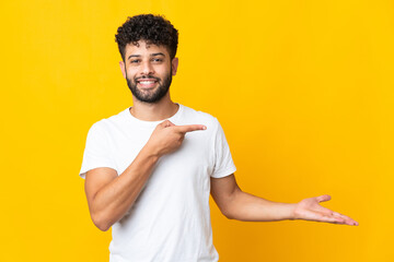Young Moroccan man isolated on yellow background holding copyspace imaginary on the palm to insert...