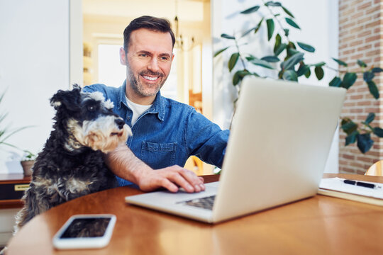 Cheerful man using laptop at home with dog on lap looking on a screen