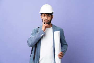 Young architect Moroccan man with helmet and holding blueprints over isolated background having doubts and thinking