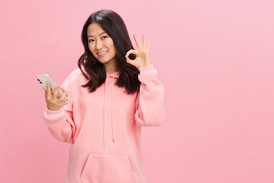 Online communication. Happy Asian student young lady in pink hoodie sweatshirt show Okay gesture posing isolated on over pink studio background. Good offer. Gadget addiction Social Media concept