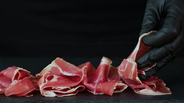 Prosciutto crudo or jamon meat with rosemary on serving board on black background. Sliced into thin pieces of ham puts on cutting board. Put jamon on serving board. Dish serving, food serving process