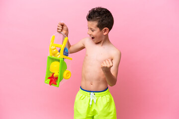 Little caucasian boy holding beach toys isolated on pink background celebrating a victory