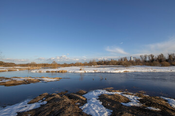 A picturesque landscape, early spring, a river with snow-covered banks, dry grass and bushes. March sunny day by the river. The first thaws, the snow is melting.