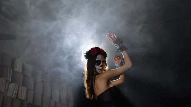 Young model with Halloween makeup in black top and lacy cuffs dances in smoke against artificial light and studio wall with books