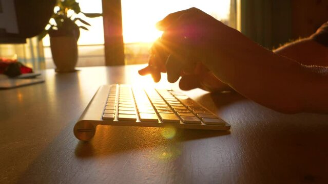 TRZIN, SLOVENIA, 20.5.2022: SLOW MOTION, CLOSE UP: Hands working and typing on computer keyboard in early morning. The rays of sunrise shining over typing fingers and wireless keyboard in home office