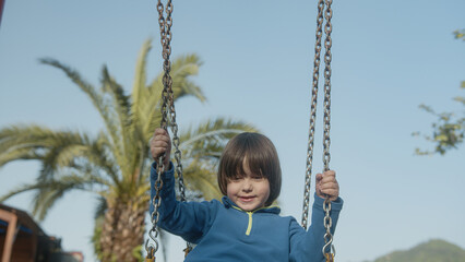 On sunny spring day, little boy or baby is smiling and having fun, swinging on the cute cheerful swing, enjoying playing in the park with his smiling cute or cute facial expressions