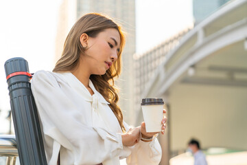 Beautiful Asian businesswoman drinking hot coffee while walking city street to working in the morning. Business woman holding coffee cup walking in busy office center district in working day rush hour