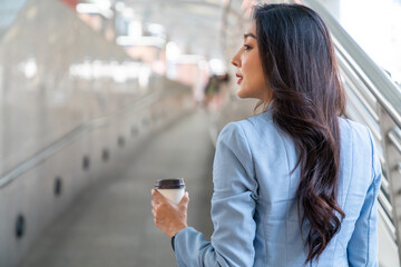 Beautiful Asian businesswoman drinking hot coffee while walking city street to working in the morning. Business woman holding coffee cup walking in busy office center district in working day rush hour
