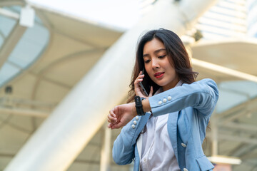 Confidence Asian businesswoman checking time on hand watch while walking in city street. Business woman working busy talking on mobile phone for online corporate business at office building district
