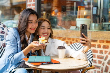 Asian woman friends using smartphone selfie together while sitting at outdoor coffee shop eating bakery and drinking coffee in urban city street. Beautiful female enjoy outdoor lifestyle in the city