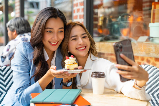 Asian woman friends using smartphone selfie together while sitting at outdoor coffee shop eating bakery and drinking coffee in urban city street. Beautiful female enjoy outdoor lifestyle in the city - Powered by Adobe