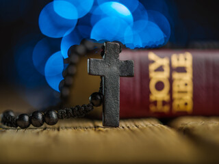 On a simple wooden table is a Catholic cross and a Holy Bible against a blue background with twinkling lights. Religion, christianity, catholicism, faith, spirituality.