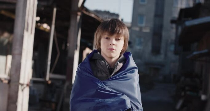 Close up portrait of little boy covered in Ukrainian flag standing outside on destroyed street looking at camera. Small kid supporting Ukraine, Russian war, conflict, crisis, patriot, patriotic