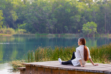 Asian woman sitting on the edge of dock with peaceful natural park during summer looking at...