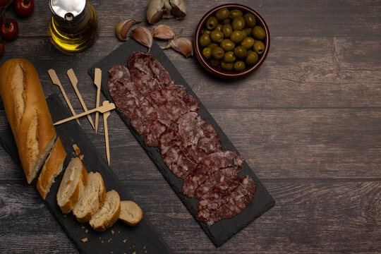 plate of cured Iberian sausage, chacina on a slate stone plate, on a wooden table with olives, garlic, olive oil and bread with space for text,