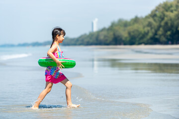 Portrait of Asian child girl in swimwear and swim ring running playing on tropical beach in sunny day. Happy little girl enjoy and fun outdoor activity lifestyle on summer travel vacation at the sea