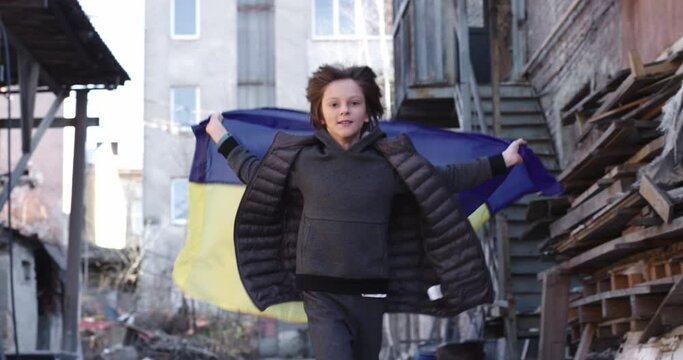Little cute boy running on street in ruined city holding Ukrainian blue and yellow flag. Small child playing in destroyed town. Child of war, conflict, crisis, Russian invasion, childhood